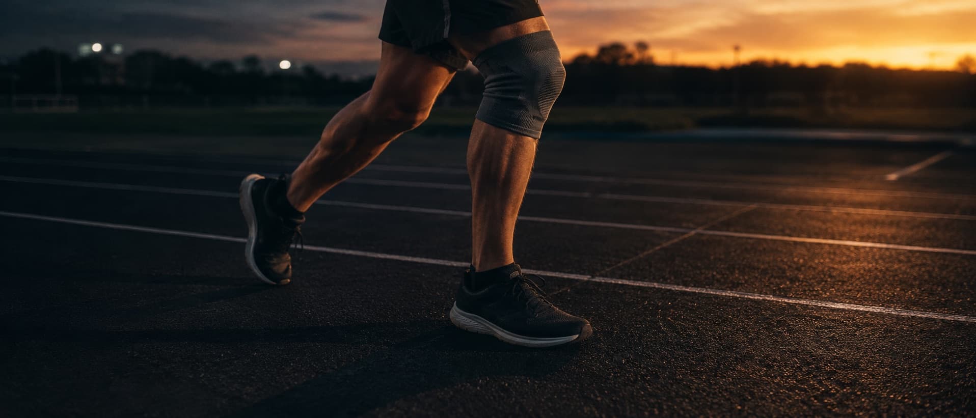 A runner's legs mid-stride on a dark track at dawn, one knee in a support sleeve